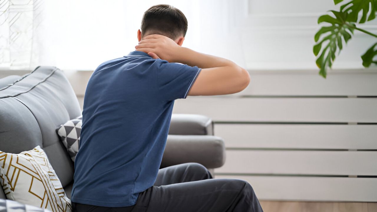 A person sits on a gray couch, turned away from the viewer, with their hand on their neck, possibly indicating discomfort related to sciatica treatment.